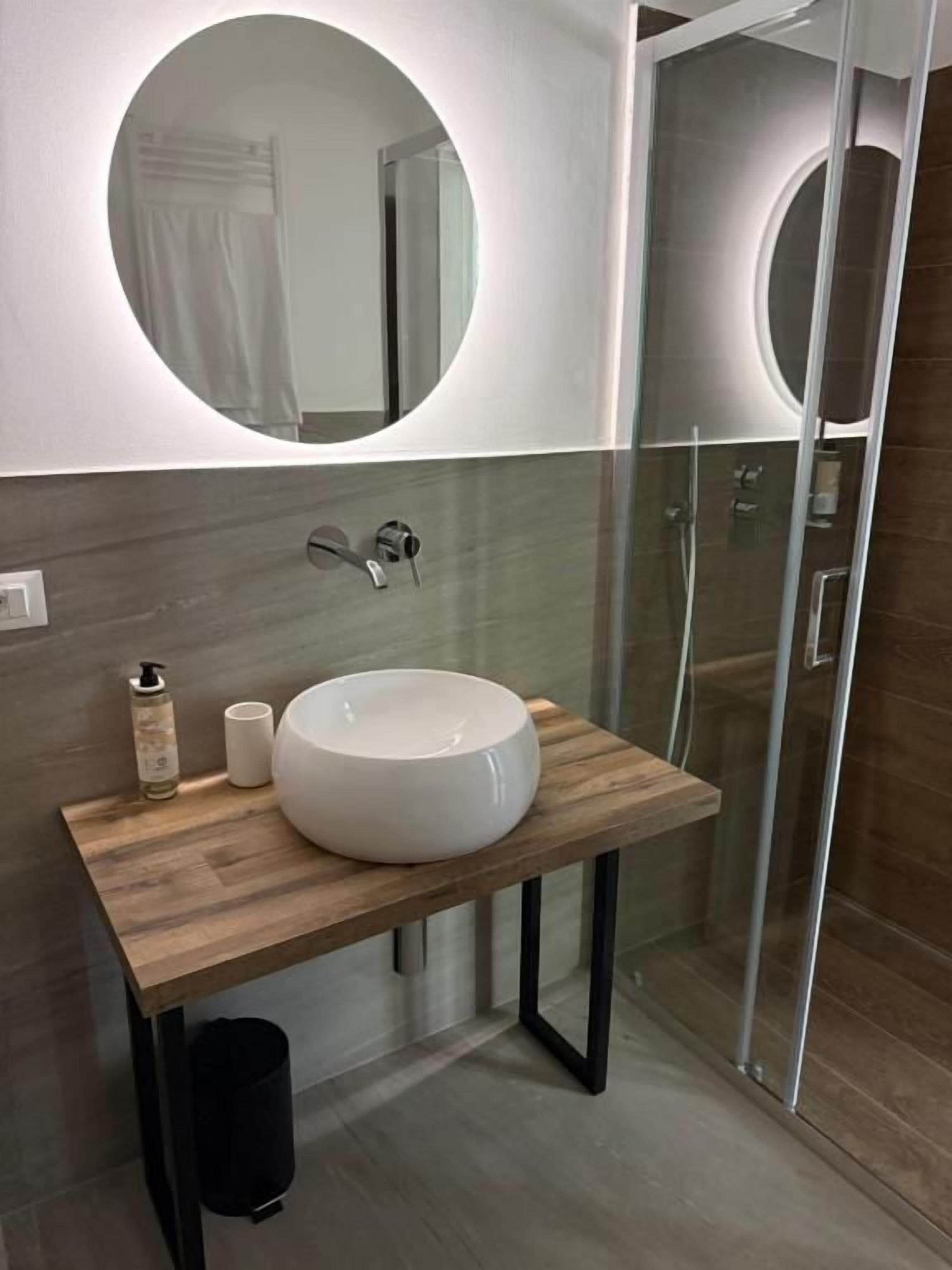 A bathroom with a round mirror, a wooden vanity, a white sink, and a glass shower enclosure.