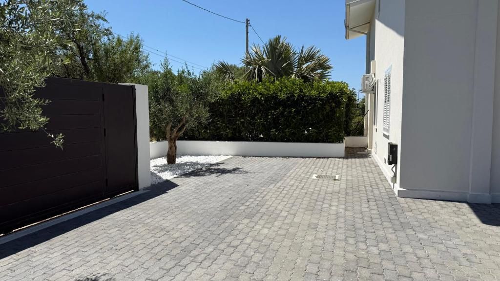 Un camino pavimentado conduce a un edificio blanco con un portón oscuro, rodeado de follaje y un cielo despejado.