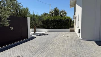Un camino pavimentado conduce a un edificio blanco con un portón oscuro, rodeado de follaje y un cielo despejado.