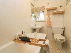 A bathroom with a bathtub, a sink, a toilet, and a wooden shelf with toiletries. - Corner Cottage (Ulverston)