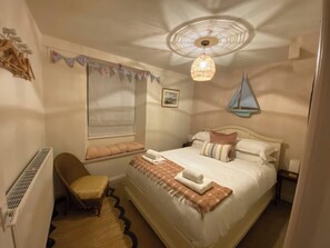 A bedroom with a bed, a chair, a window with blinds, a wall-mounted sailboat decoration, and a ceiling light fixture. - Corner Cottage (Ulverston)