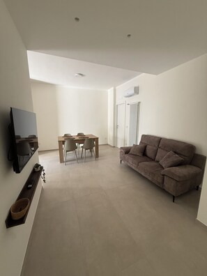 A modern living room with a flat-screen TV mounted on the wall, a brown sofa, a dining table with chairs, and a ceiling light fixture. - Quaint 2-bedroom apartment with AC in charming Bari (Bari)