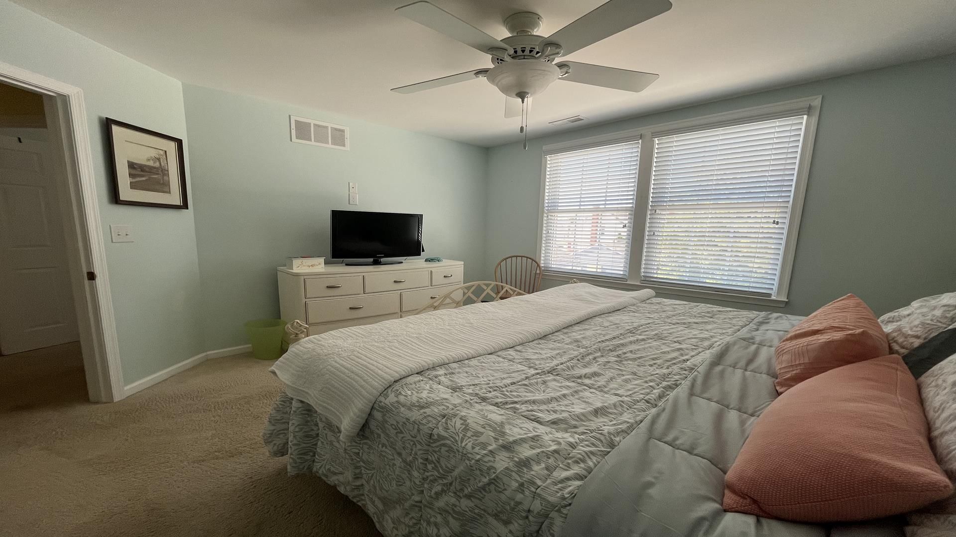 A bedroom with a bed, a television on a dresser, a ceiling fan, and a window with blinds.
