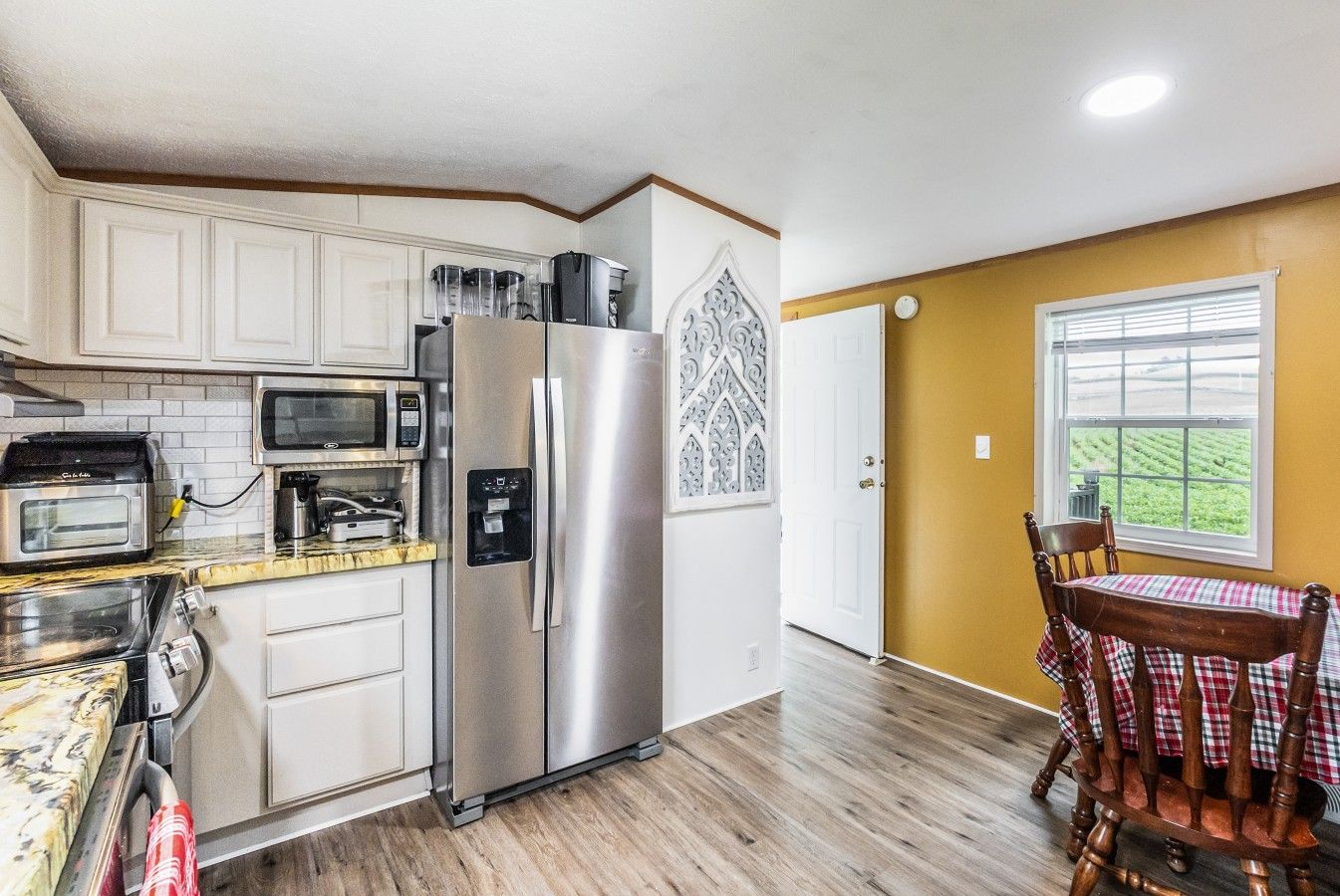 A compact kitchen with a stainless steel refrigerator, microwave, and toaster, a wooden dining table with a checkered tablecloth, and a window with a view of a green field.