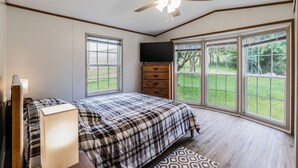 Ein Schlafzimmer mit Bett, Kommode, Fernseher und großen Fenstern mit Blick ins Freie.