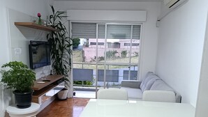 A modern living room with a white sofa, a dining table with chairs, a TV, and a potted plant.