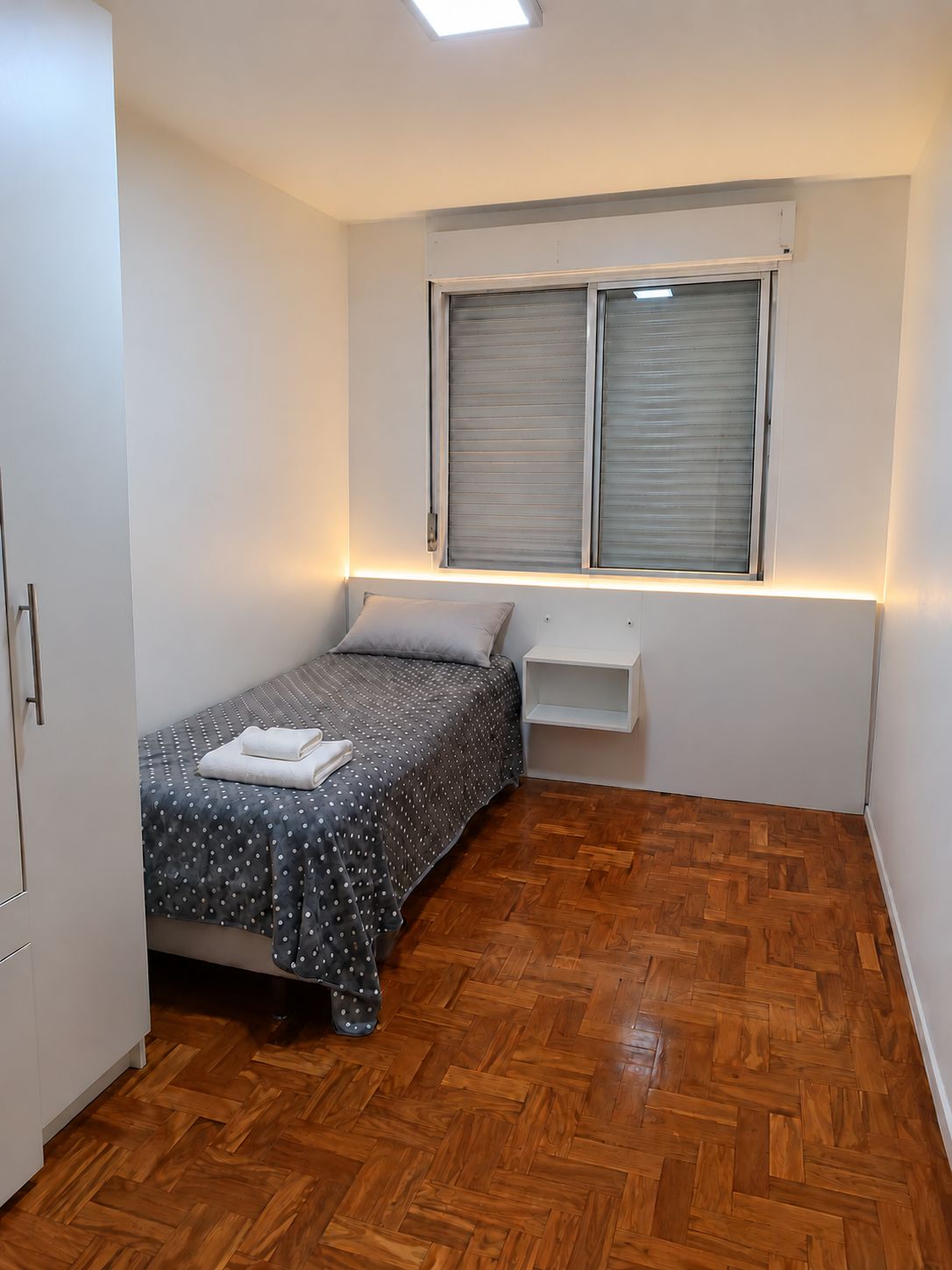 Un dormitorio pequeño y ordenado con cama, mesita de noche, armario y ventana con persianas.