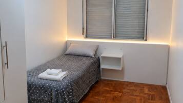 Un dormitorio pequeño y ordenado con cama, mesita de noche, armario y ventana con persianas.