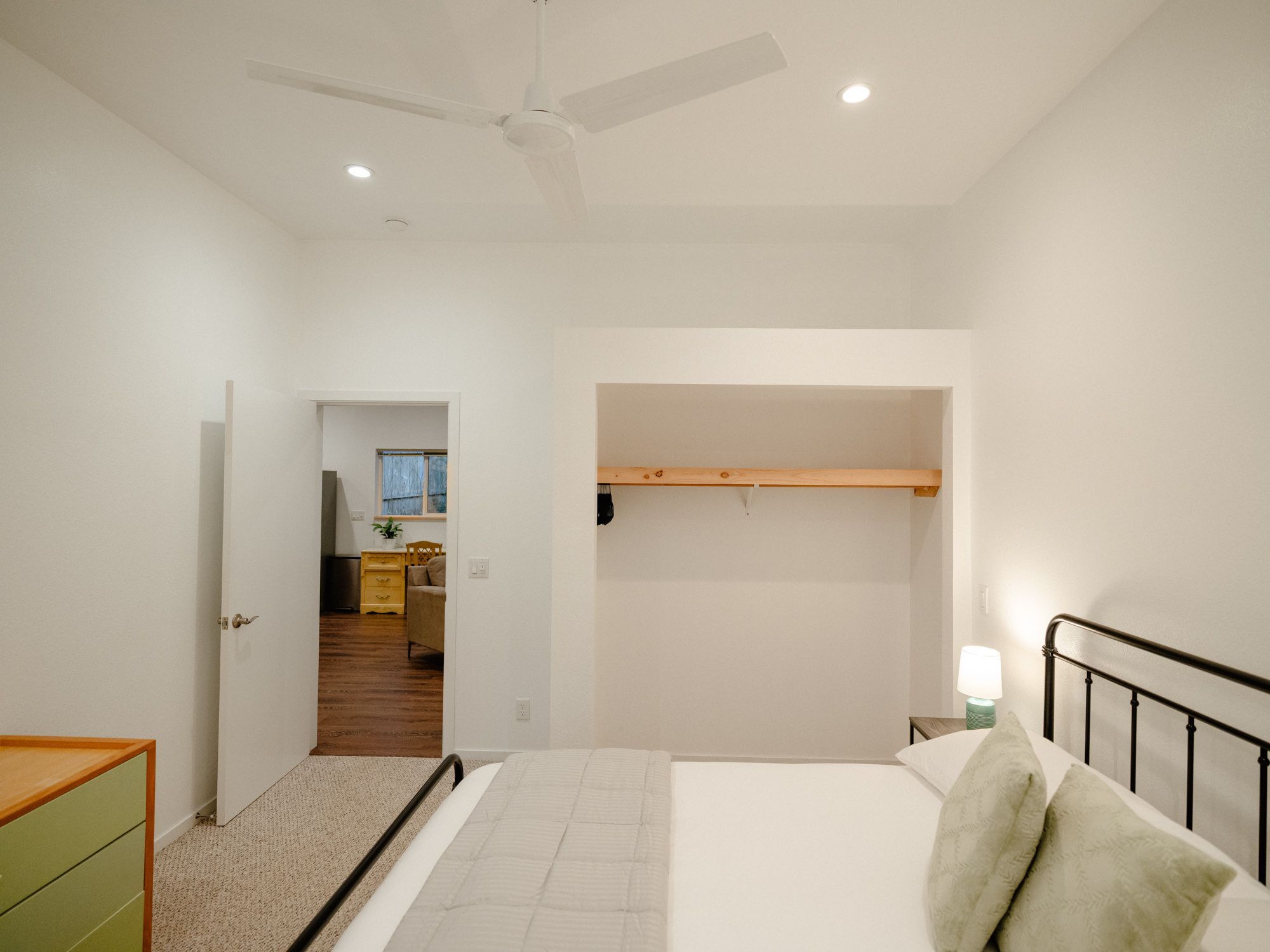 Une chambre à coucher avec un lit, une table de chevet avec une lampe, un ventilateur de plafond et une étagère en bois.