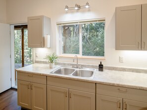A kitchen with a double sink, a window with a view of trees, and beige cabinetry. - The Nook | Modern Redwood Escape Near Gualala (Gualala)
