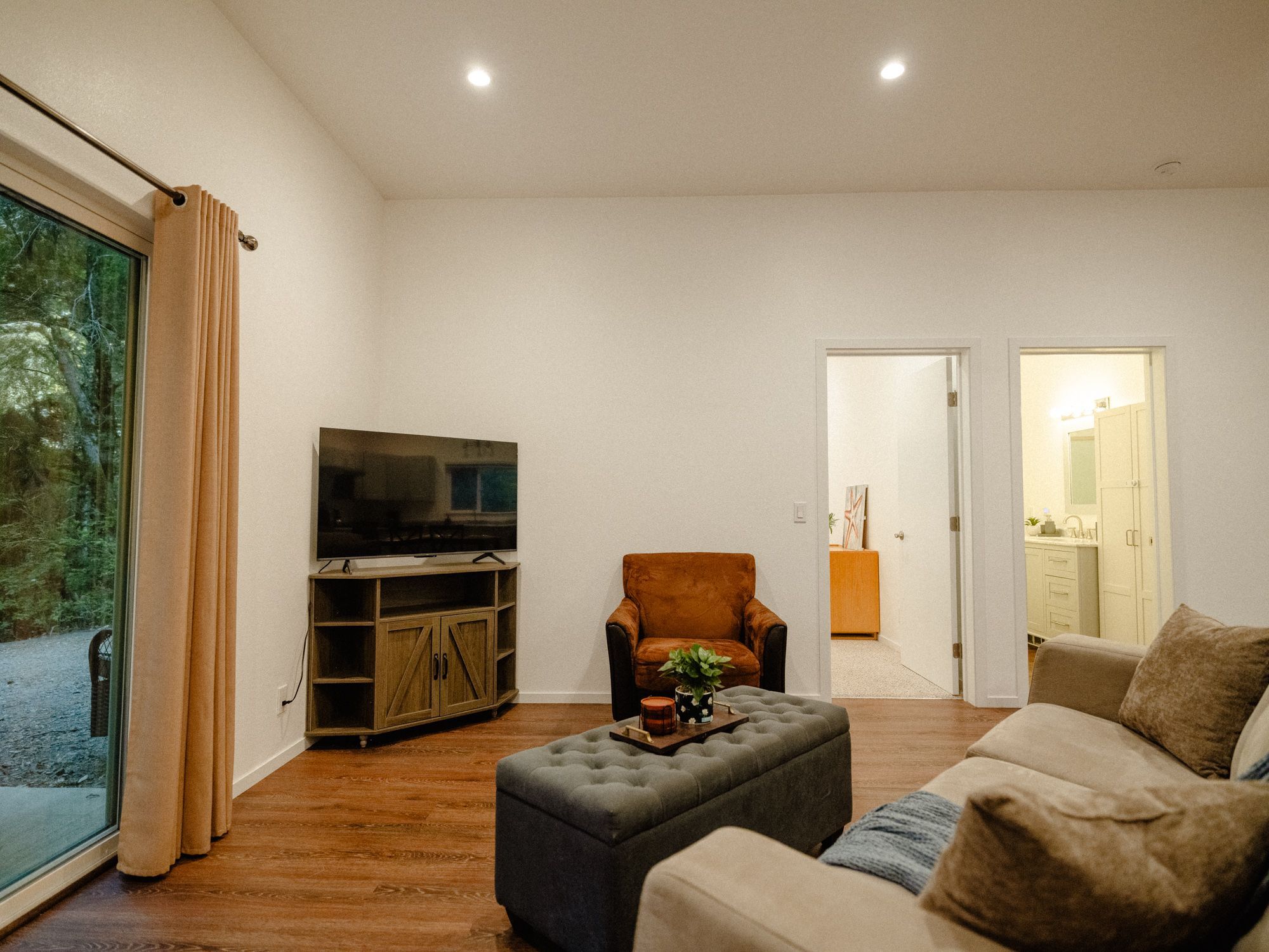 A living room with a television, a sofa, an ottoman, and a chair. There is a sliding glass door leading to a balcony.