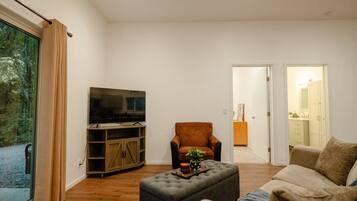 A living room with a television, a sofa, an ottoman, and a chair. There is a sliding glass door leading to a balcony.