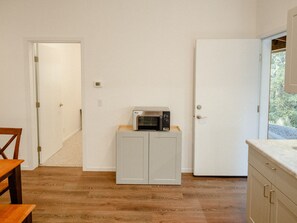 A compact kitchen with a microwave on a small cabinet, a wooden dining table, and a view through an open door. - The Nook | Modern Redwood Escape Near Gualala (Gualala)
