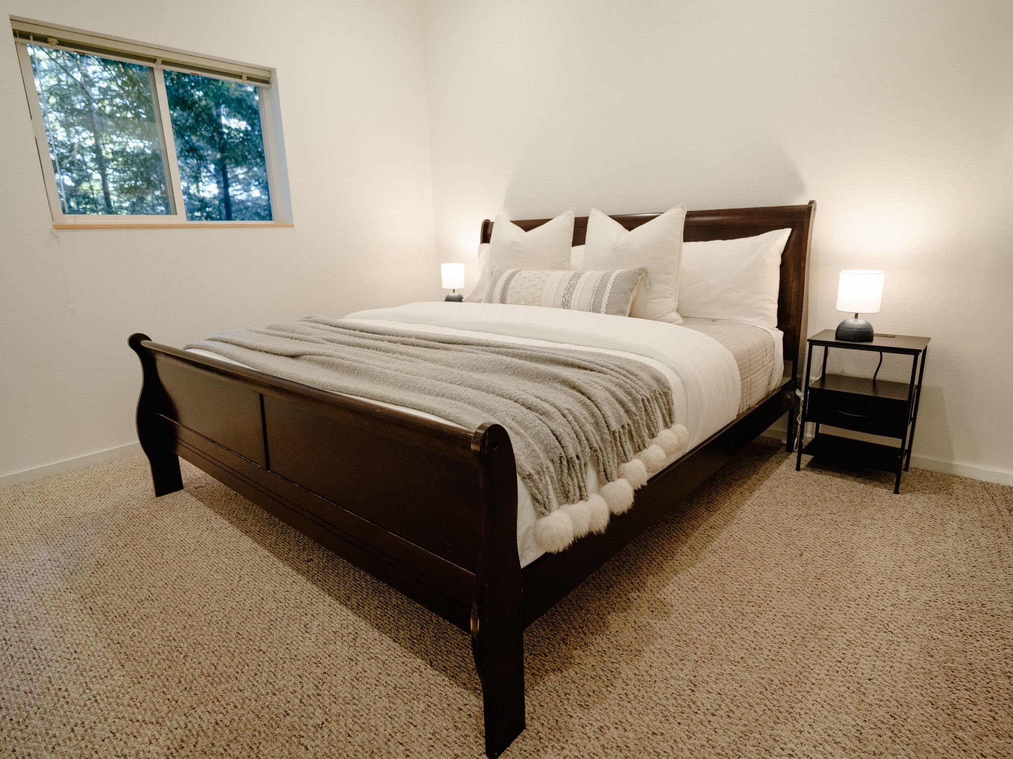 A bedroom with a wooden bed, white bedding, a nightstand with a lamp, and a window with a view of trees.