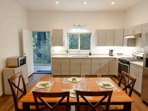 A modern kitchen with a dining area, featuring a large window, stainless steel appliances, and a wooden table set for two. - The Nook | Modern Redwood Escape Near Gualala (Gualala)