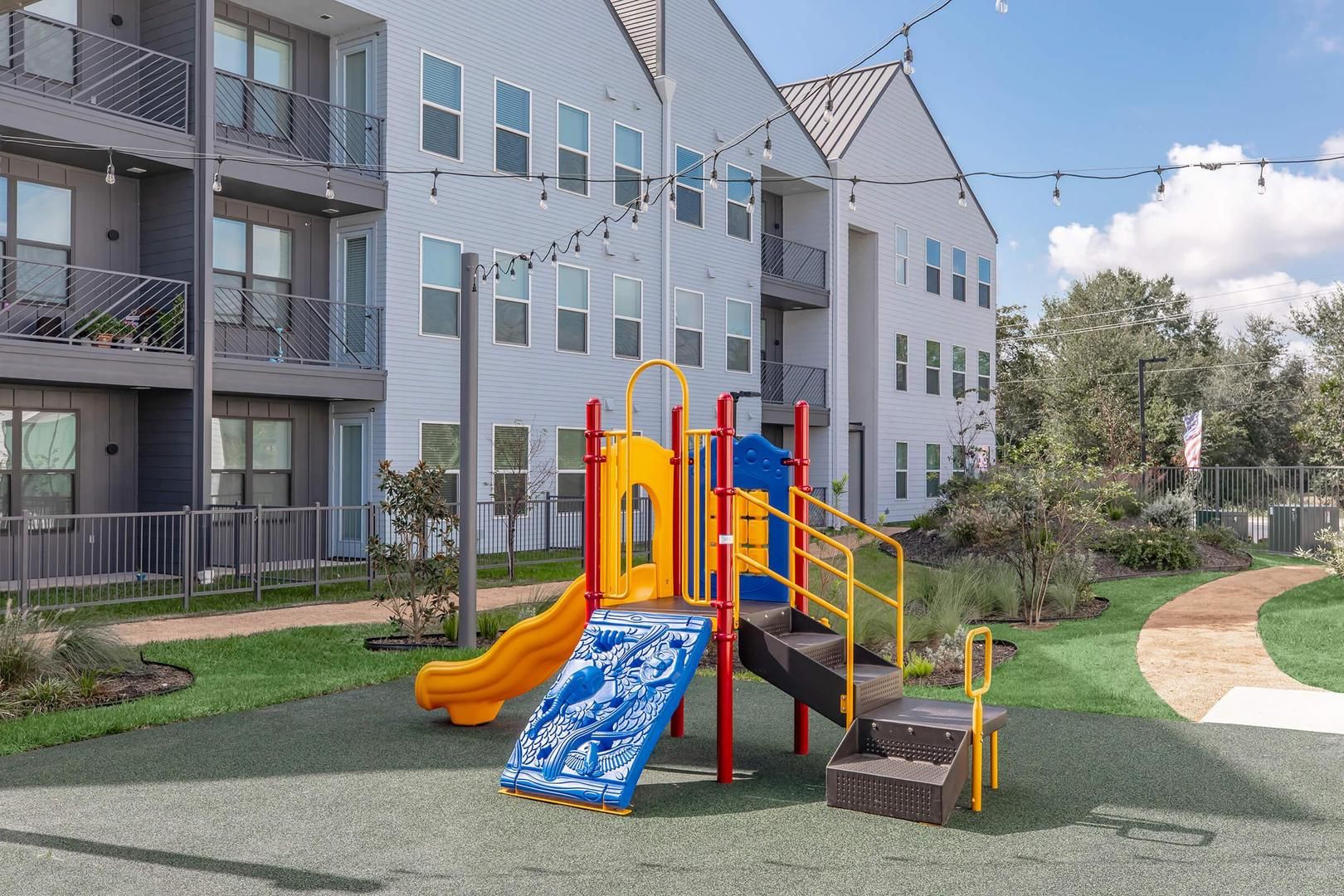 Ein Spielplatz mit Rutsche und Klettergerüsten, umgeben von einem gepflegten Rasen und einem mehrstöckigen Gebäude im Hintergrund.