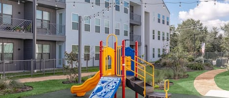 Un parque infantil con tobogán y estructuras para escalar, rodeado de césped cuidado y un edificio de varios pisos al fondo.