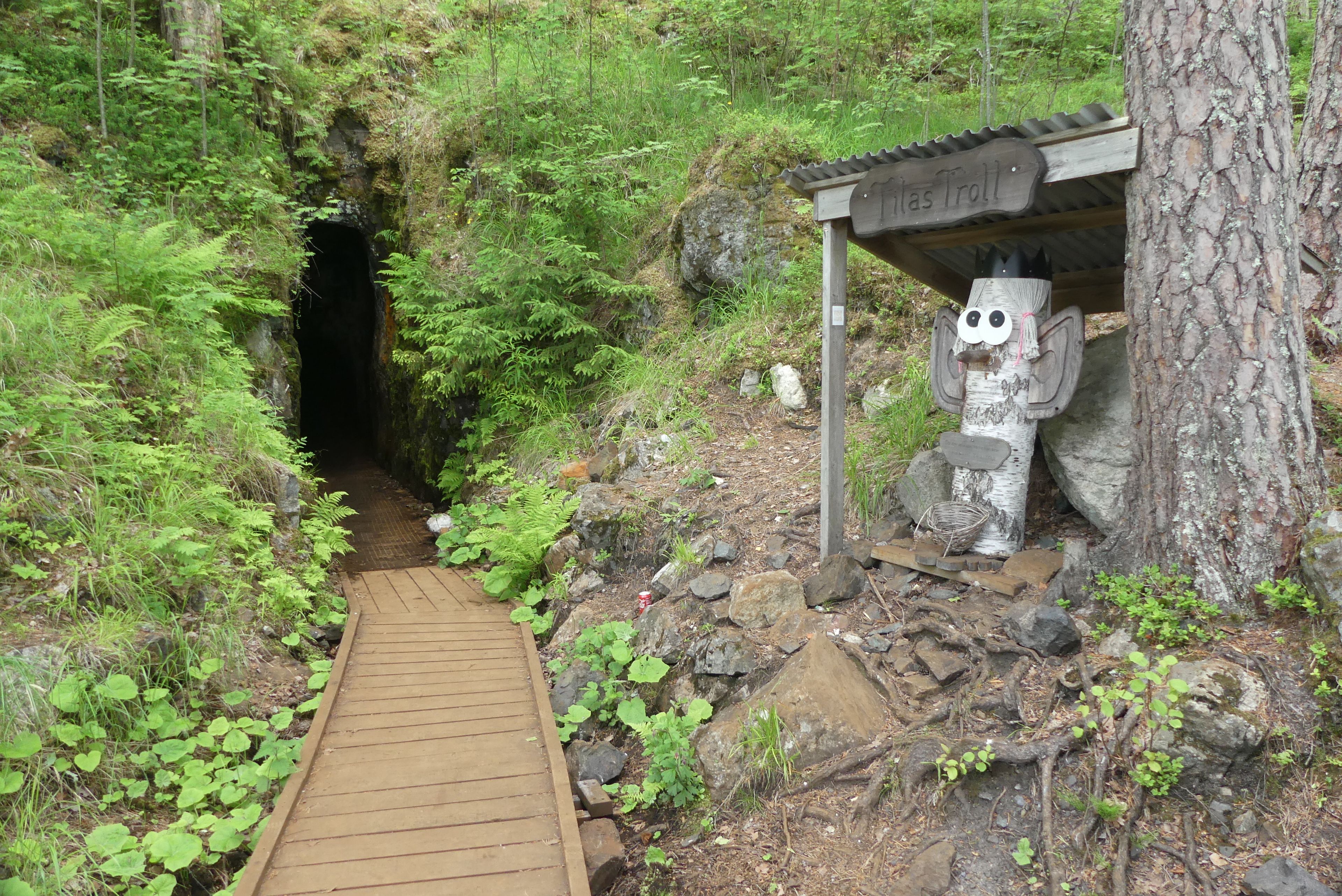 A narrow wooden pathway leading to a tunnel, surrounded by lush greenery and a wooden structure with a sign reading 'Las Troll'.