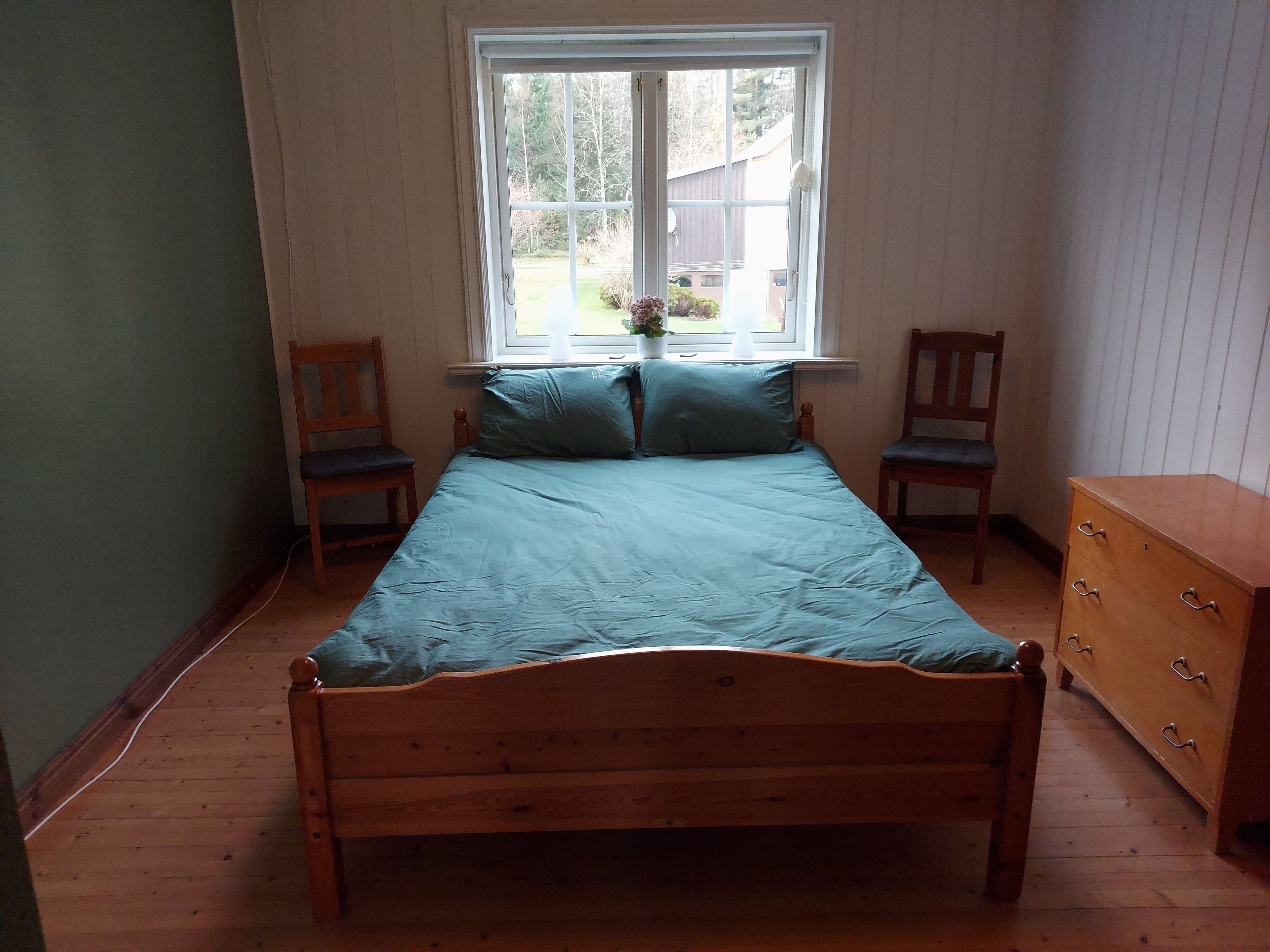 A single-bed room with a wooden headboard, two wooden chairs, a wooden dresser, and a window with a view of greenery outside.