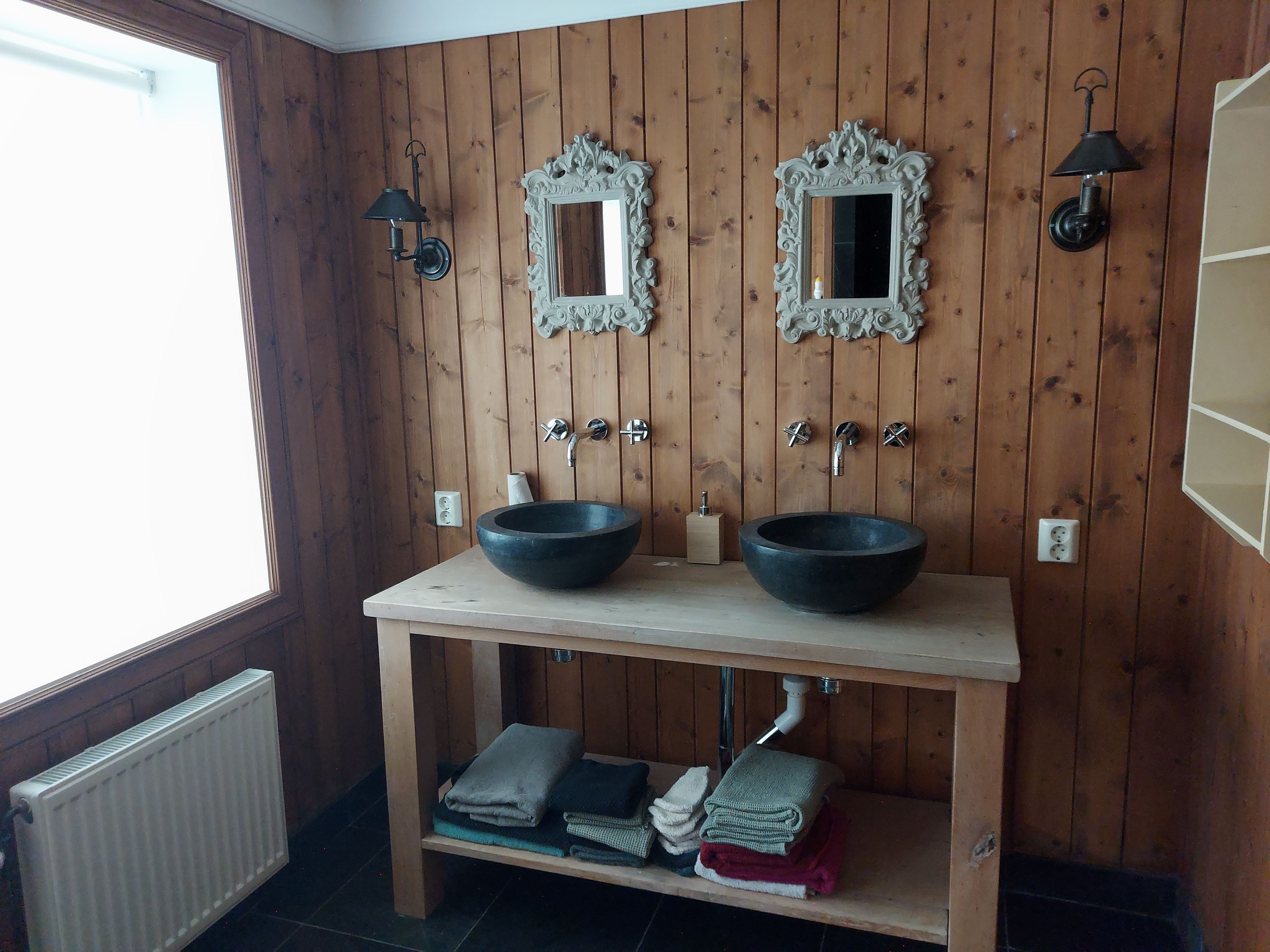Un baño con paredes de madera panelada, dos espejos, un lavamanos doble y un estante con toallas dobladas.