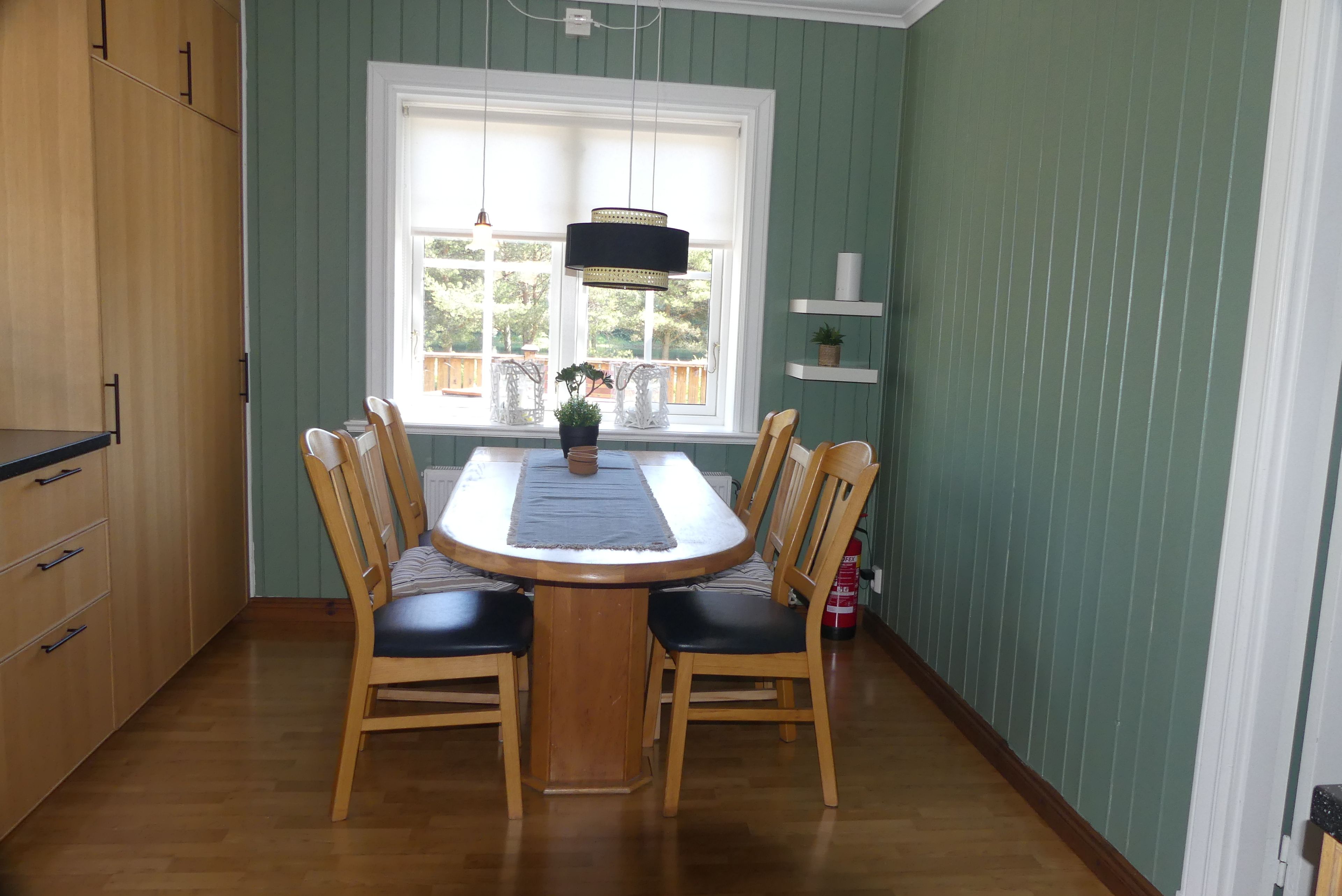 Uma sala de jantar com uma mesa redonda, quatro cadeiras, uma janela com vista para o exterior, uma luminária pendente e um extintor de incêndio na parede.