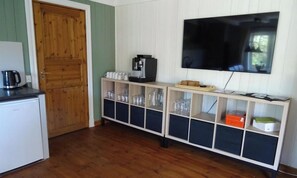 A room with a wooden door, a coffee machine, a TV mounted on the wall, and a cabinet with glassware.