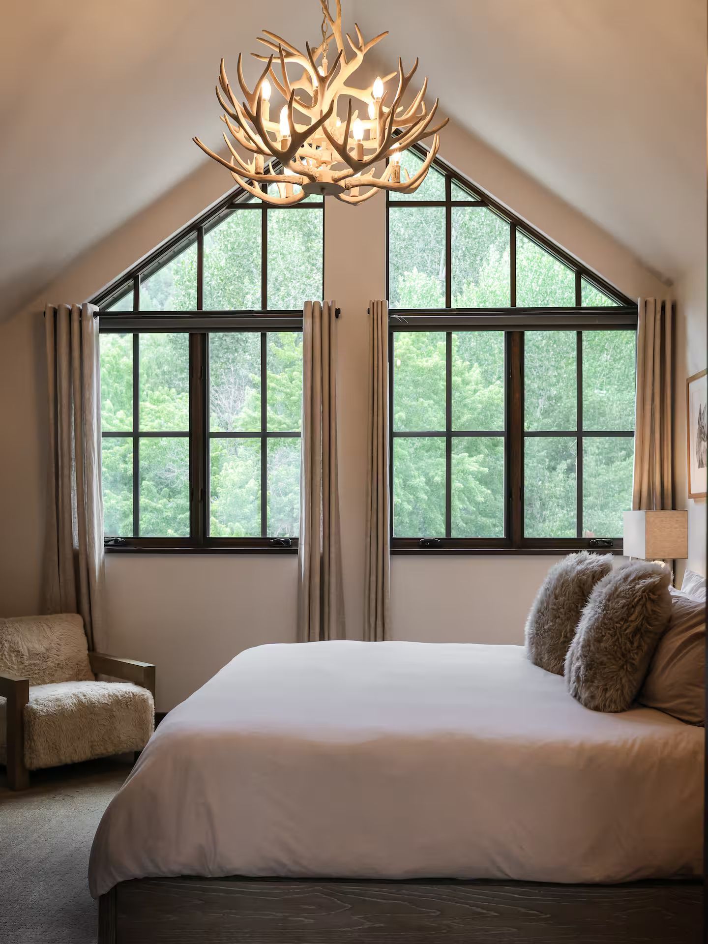 A bedroom with a large bed, a chandelier, and a view of trees through the windows.