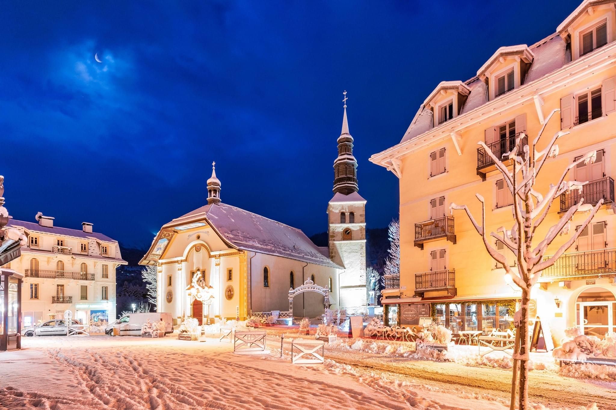 A snow-covered street with a church and adjacent buildings, including a hotel with balconies.