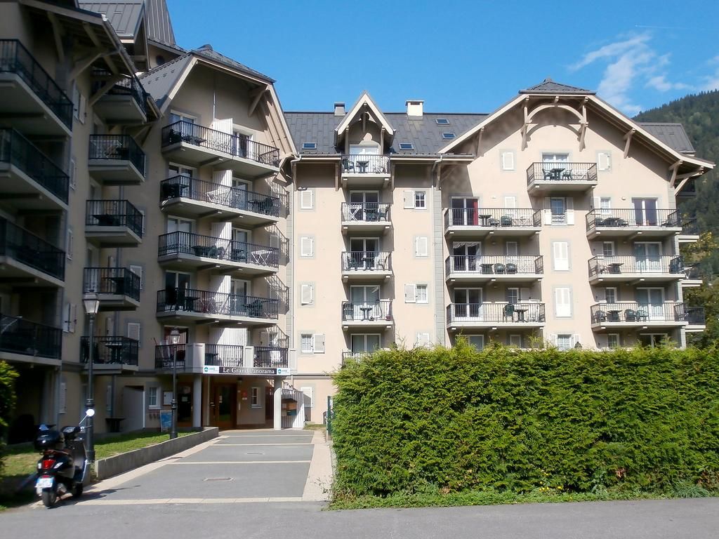 A multi-story residential building with balconies, a parking area, and a hedge in the foreground.