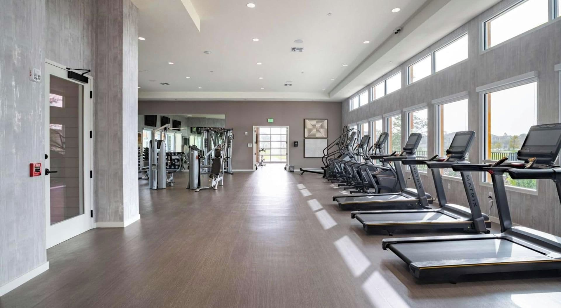 Ein Fitnessstudio mit Laufbändern, Kraftgeräten und einer großen Wand mit Fenstern.