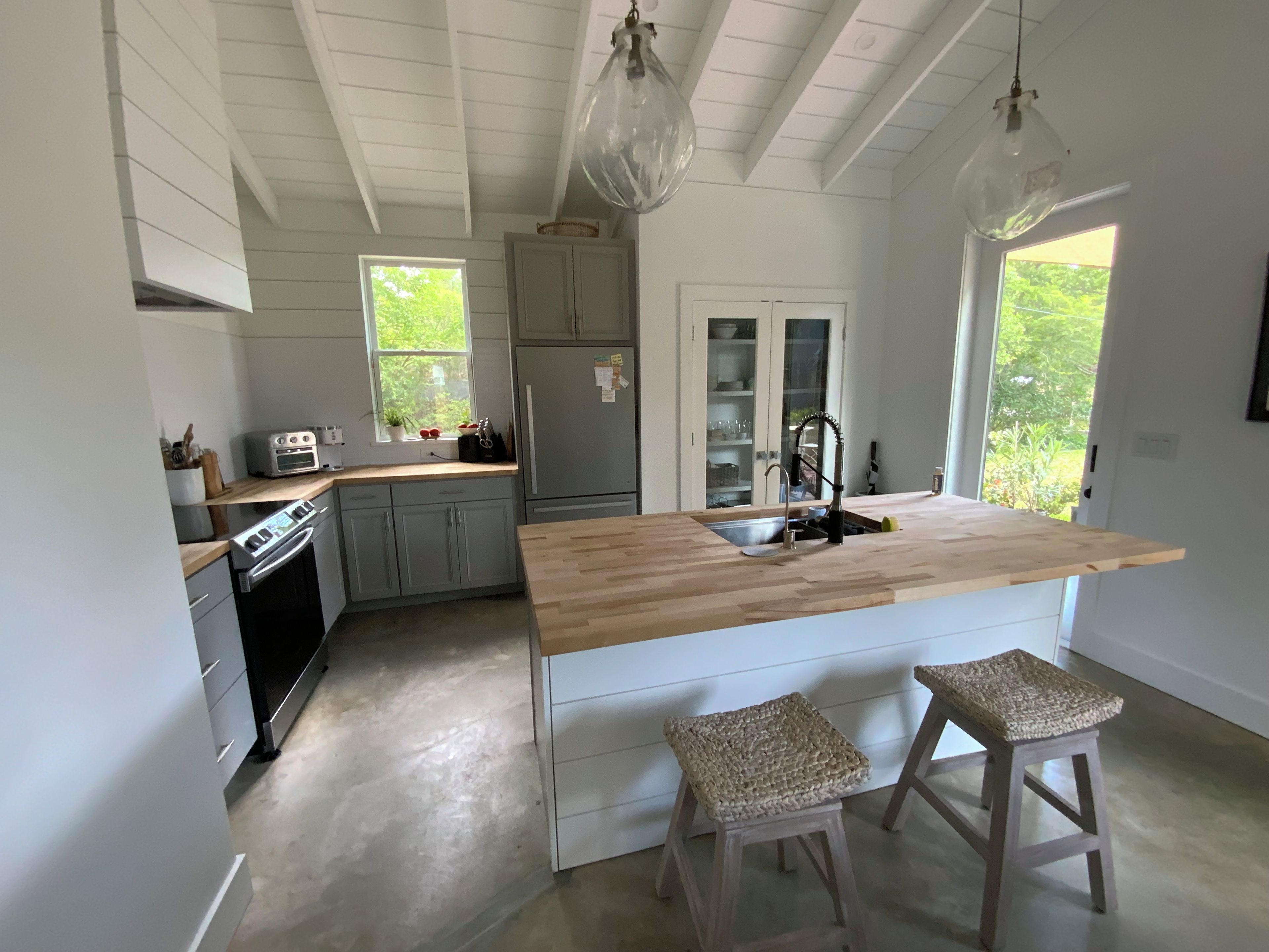 A kitchen with a wooden island, two stools, a refrigerator, and a window with a view of greenery.