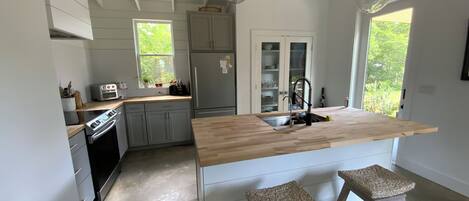 A kitchen with a wooden island, two stools, a refrigerator, and a window with a view of greenery.