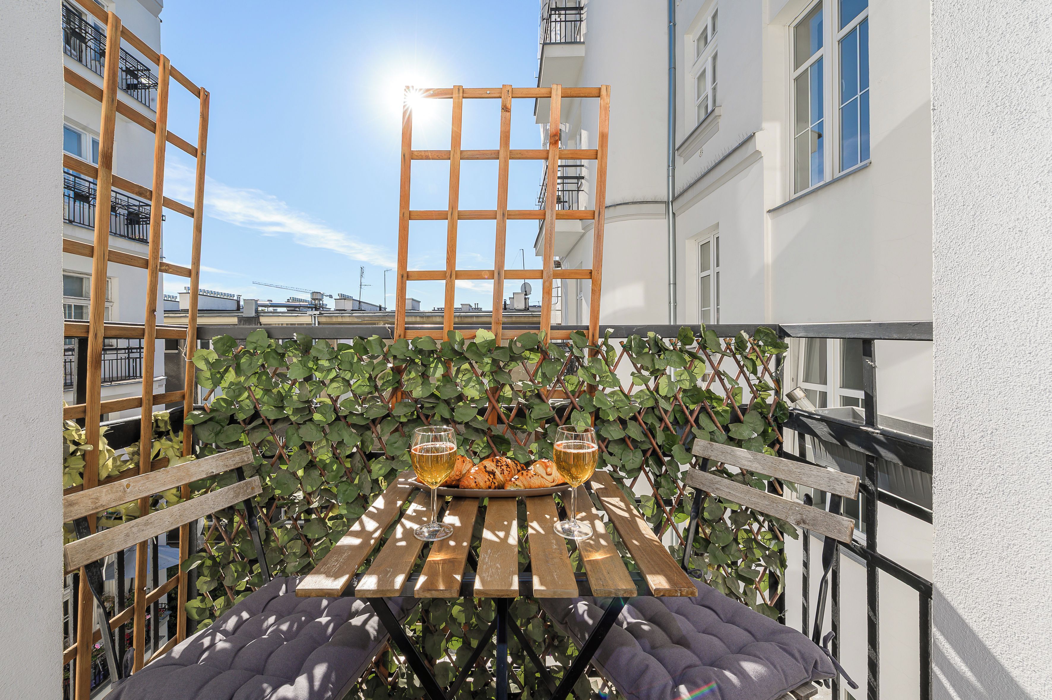 A wooden table with a glass of beer and croissants, set against a backdrop of a white building and a clear sky.