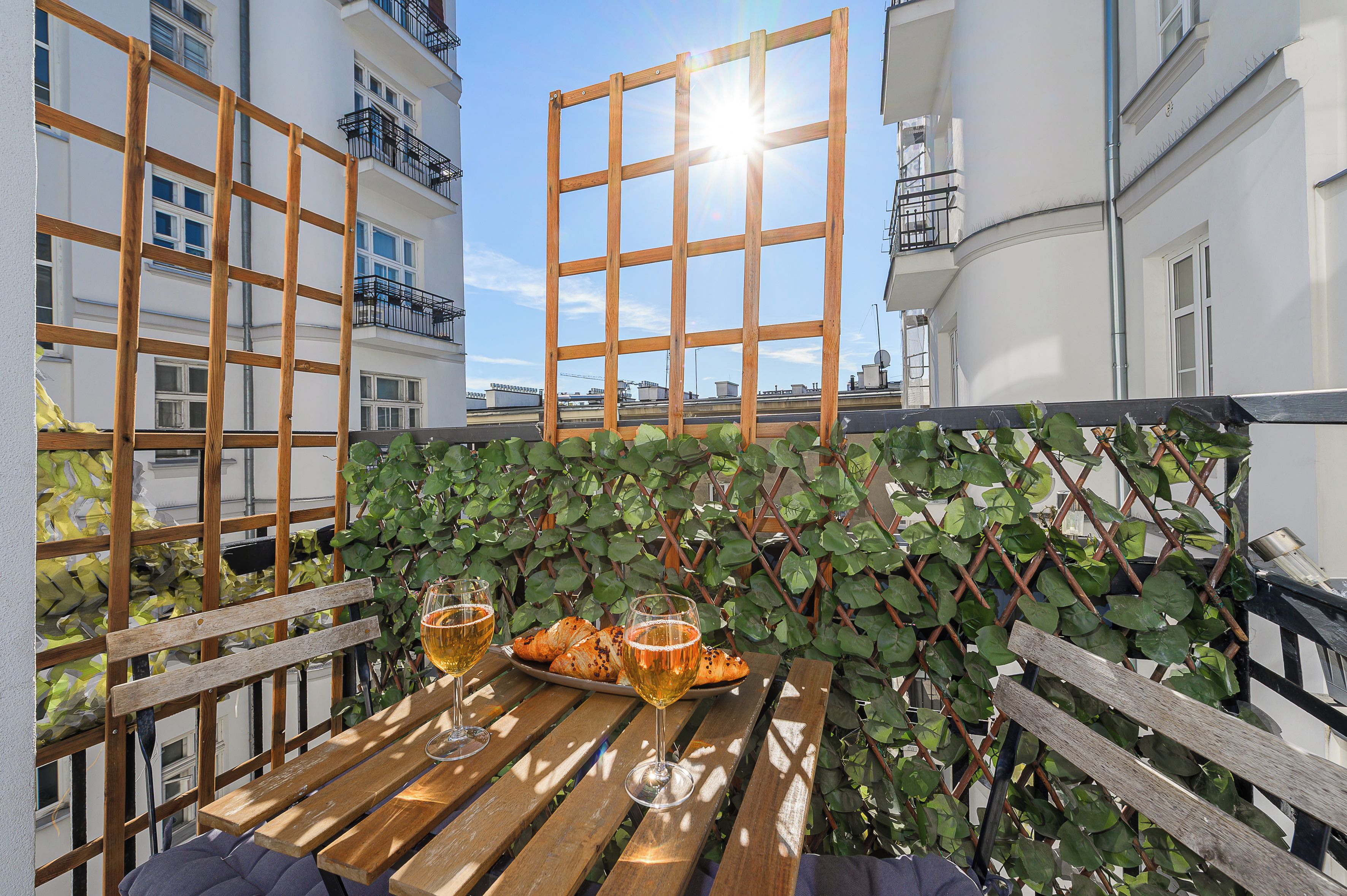 A wooden table with two glasses of beer and a plate of croissants, set against a backdrop of a white building with balconies and a clear sky.