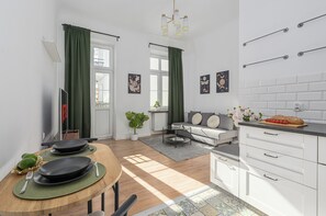 Eine moderne Küche mit weißen Schränken, eine Essecke mit einem runden Tisch und ein Wohnbereich mit einer Couch und einer Pflanze.