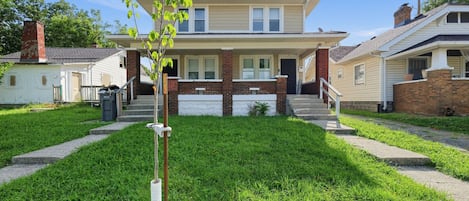 A two-story residential building with a front porch, a brick chimney, and a small tree planted in the front yard.