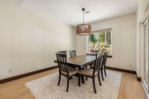 Een eethoek met een houten tafel, stoelen, een vloerkleed en een raam met uitzicht op bomen.