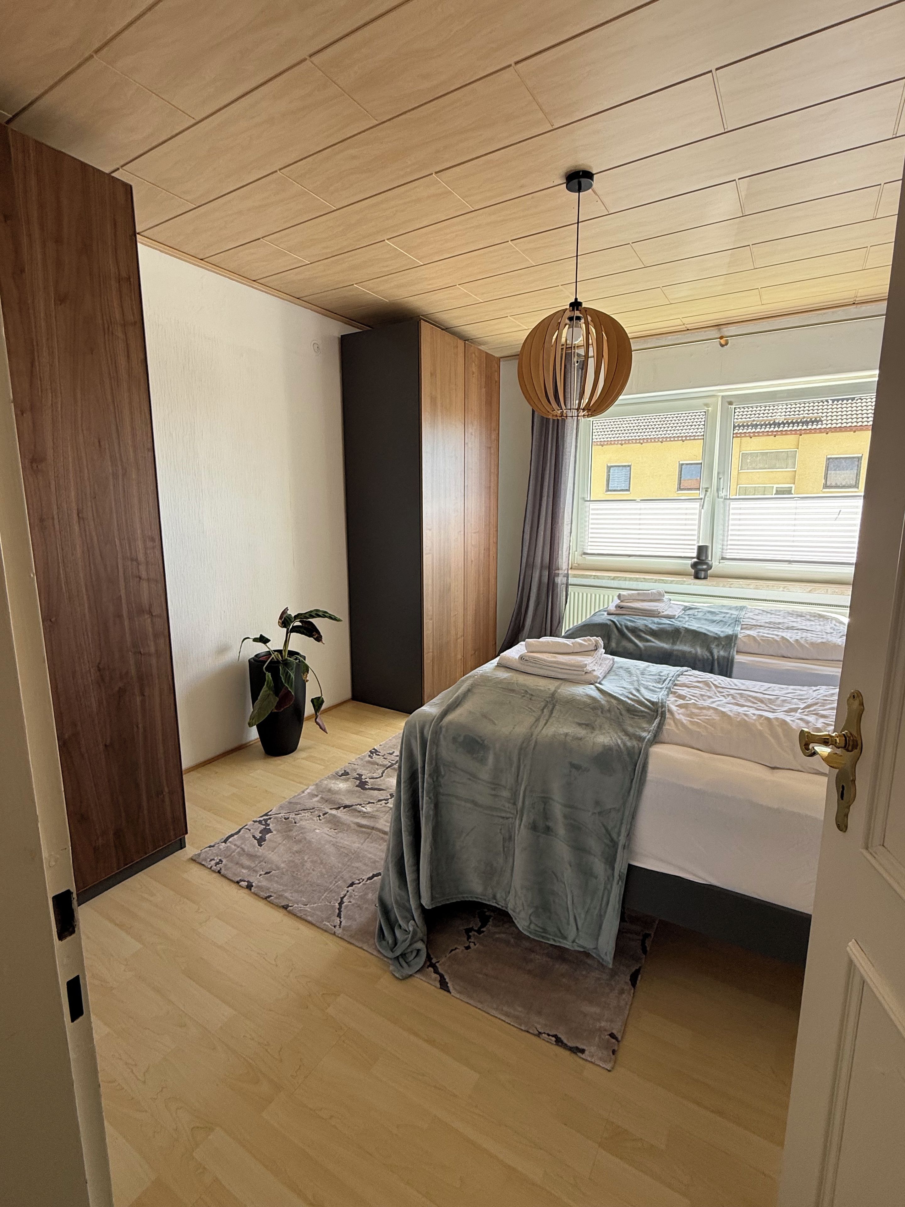 Ein Schlafzimmer mit einem Bett, einer Hängelampe, einem Blumentopf und Blick ins Freie.