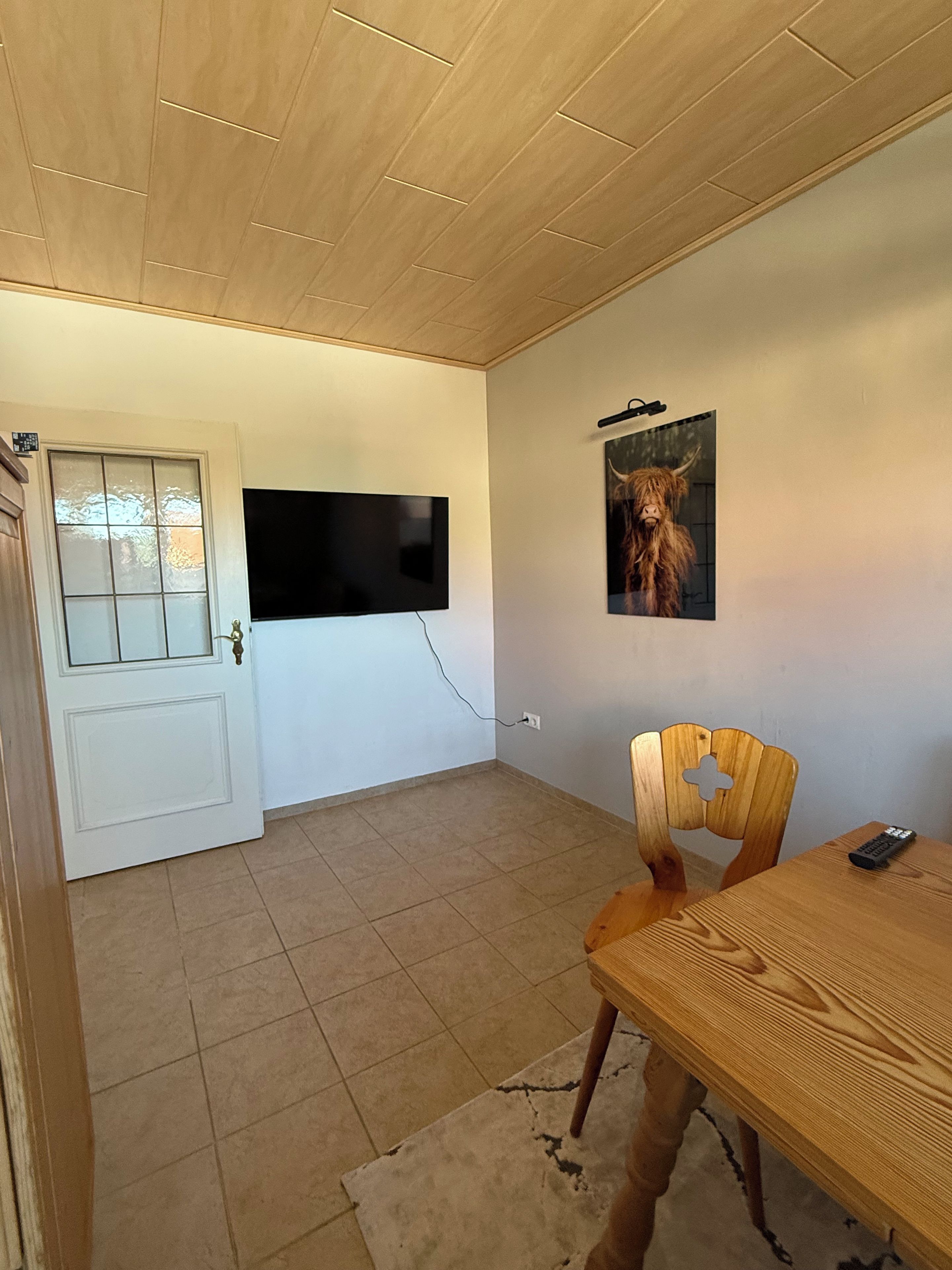 Ein Zimmer mit Holzboden, einem an der Wand befestigten Flachbildfernseher, einem Holzes Esstisch mit Stuhl und einer Tür mit Glasfenster.