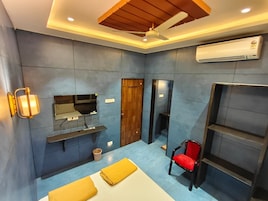 Quarto de hotel pequeno, com cama, televisão, guarda-roupa, cadeira e prateleira.