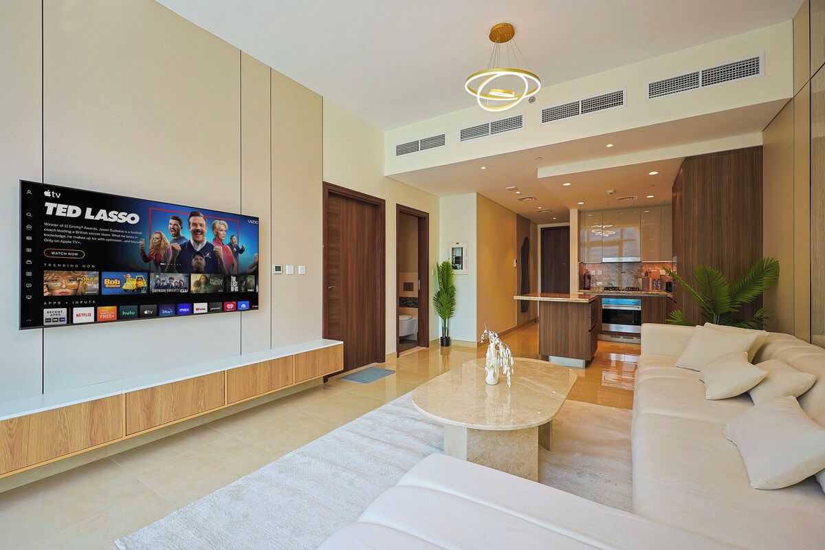 A modern living room with a flat-screen TV, a sofa, a coffee table, and a kitchen area in the background.
