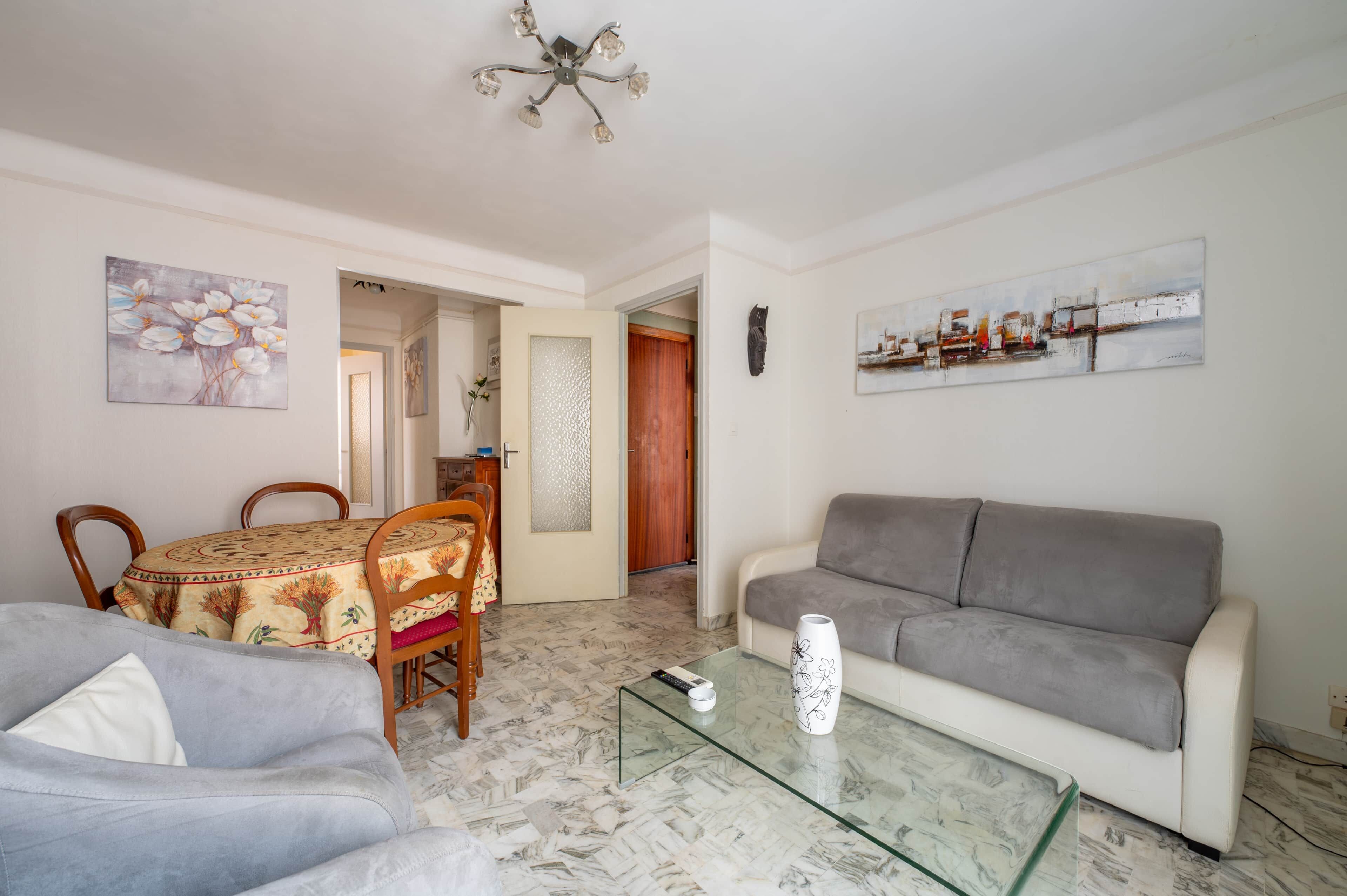 Una sala de estar con un sofá gris, una mesa de centro de vidrio, una mesa de comedor con sillas y obras de arte en las paredes.
