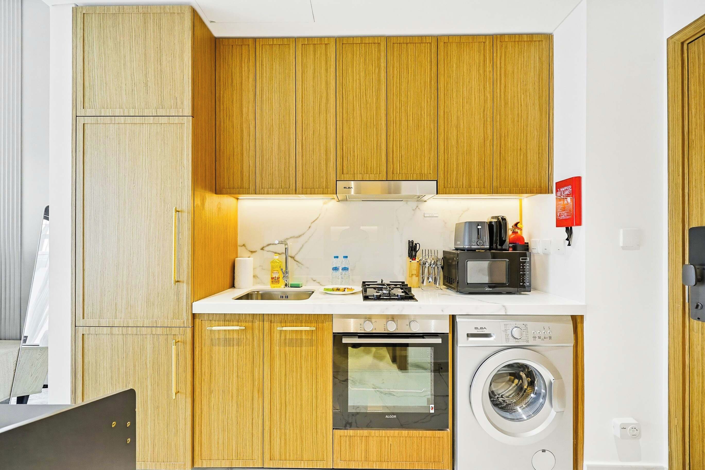 Een kleine keuken met houten kastjes, een ingebouwde magnetron, een fornuis en een wasmachine.