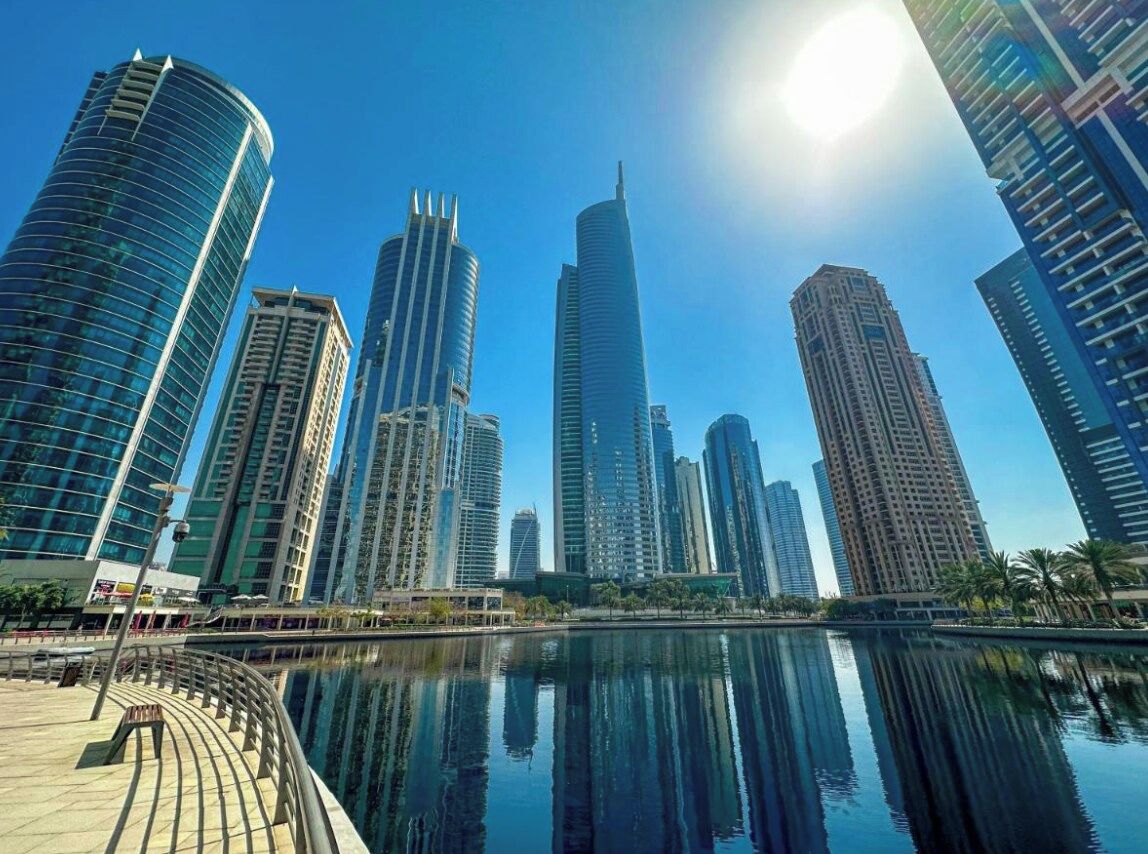 Un paysage urbain avec des immeubles modernes de grande hauteur, une étendue d’eau calme qui reflète les bâtiments, et un ciel dégagé.