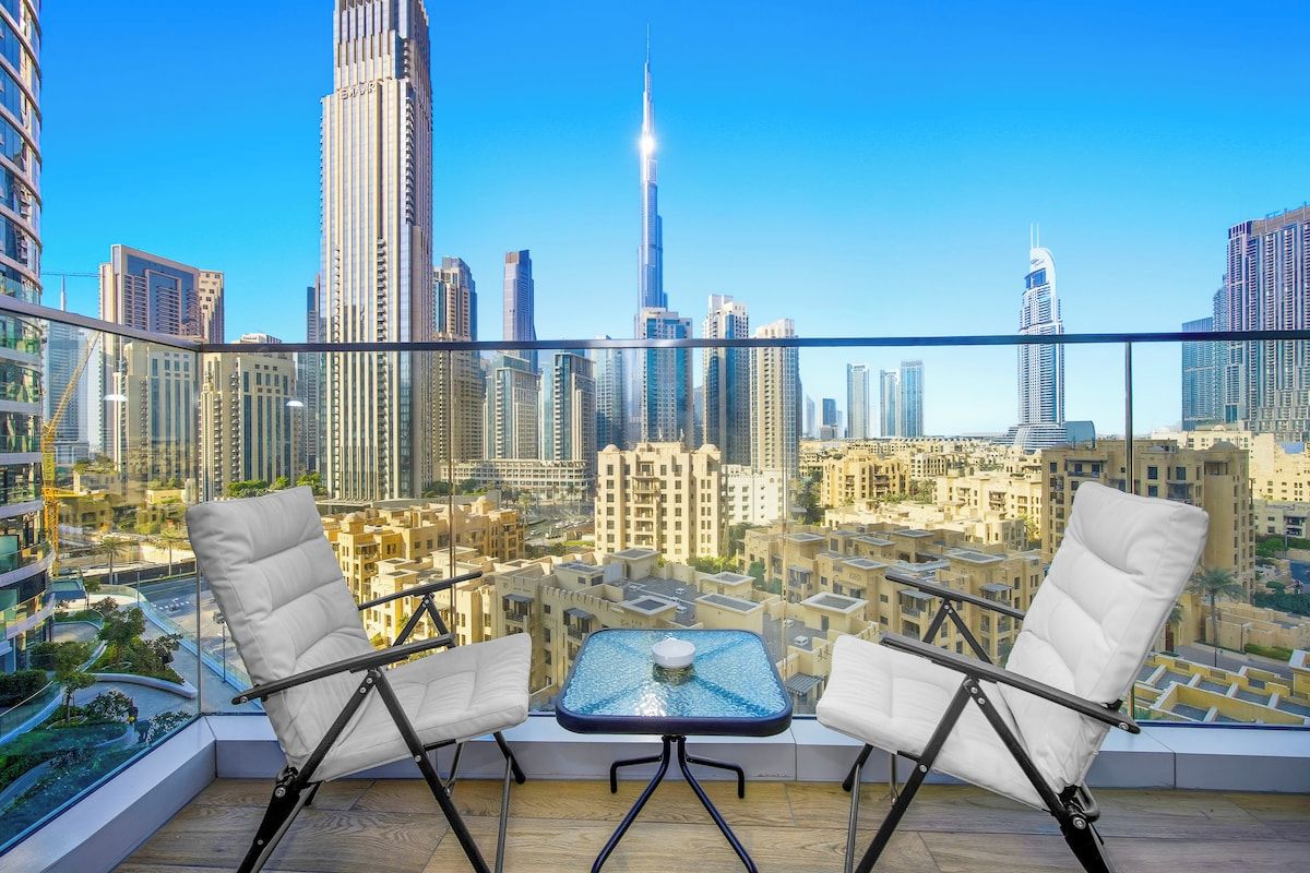 A balcony with two white cushioned chairs and a small table, overlooking a cityscape with high-rise buildings.