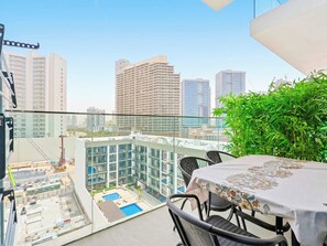 Ein Balkon mit Glasgeländer, der auf eine Stadtlandschaft blickt, ein Tisch mit einer Tischdecke mit Blumenmuster und ein kleiner Poolbereich.