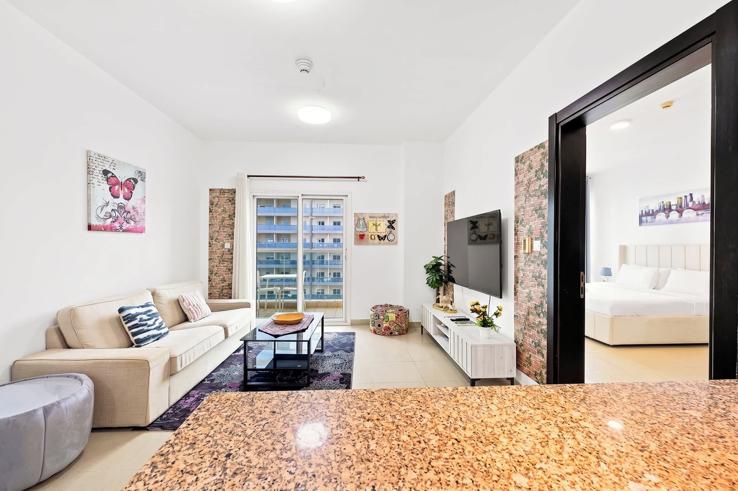 Ein modernes Wohnzimmer mit einer Couch, einem Couchtisch und einem Fernseher. Es gibt einen Balkon mit Blick auf ein Gebäude.
