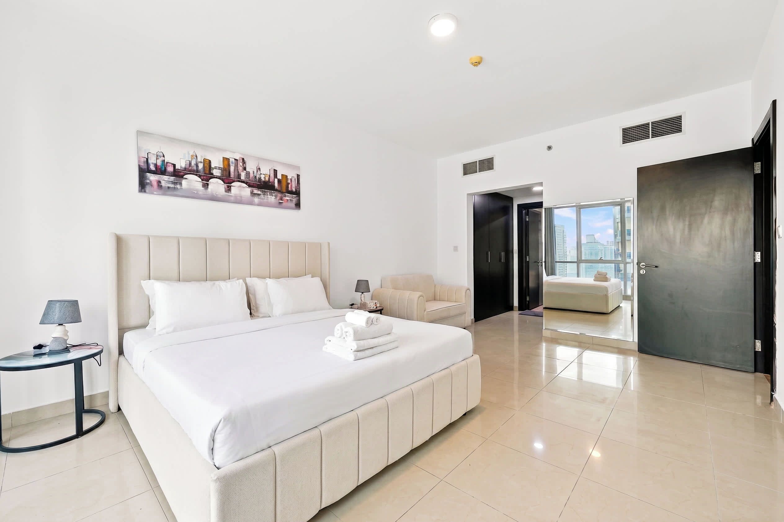 A modern hotel room with a large bed, a seating area, a cityscape painting, and a view through a door.