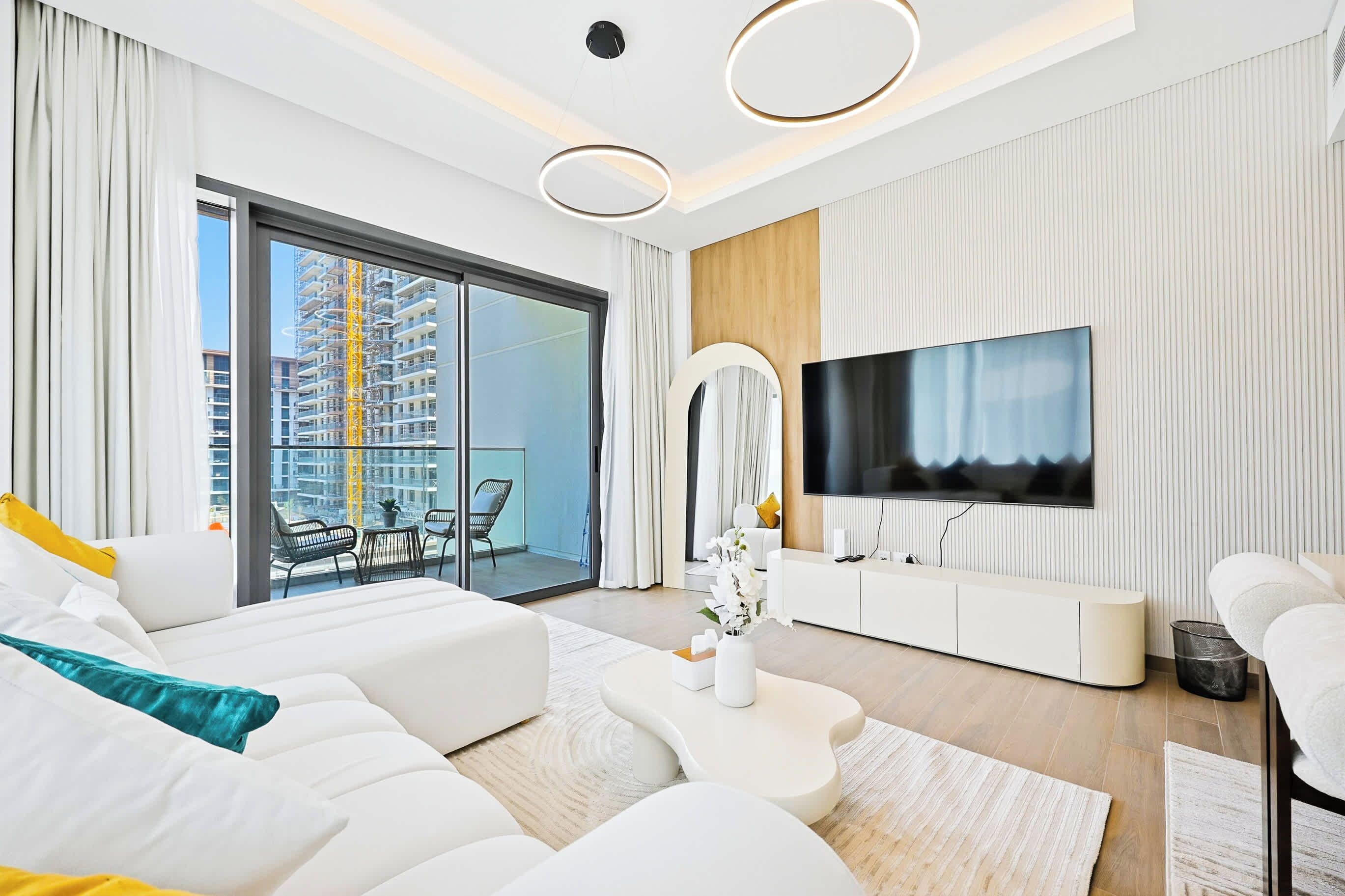 A modern living room with a large flat-screen TV, white sofas, a coffee table, and a balcony with a view of a construction site.
