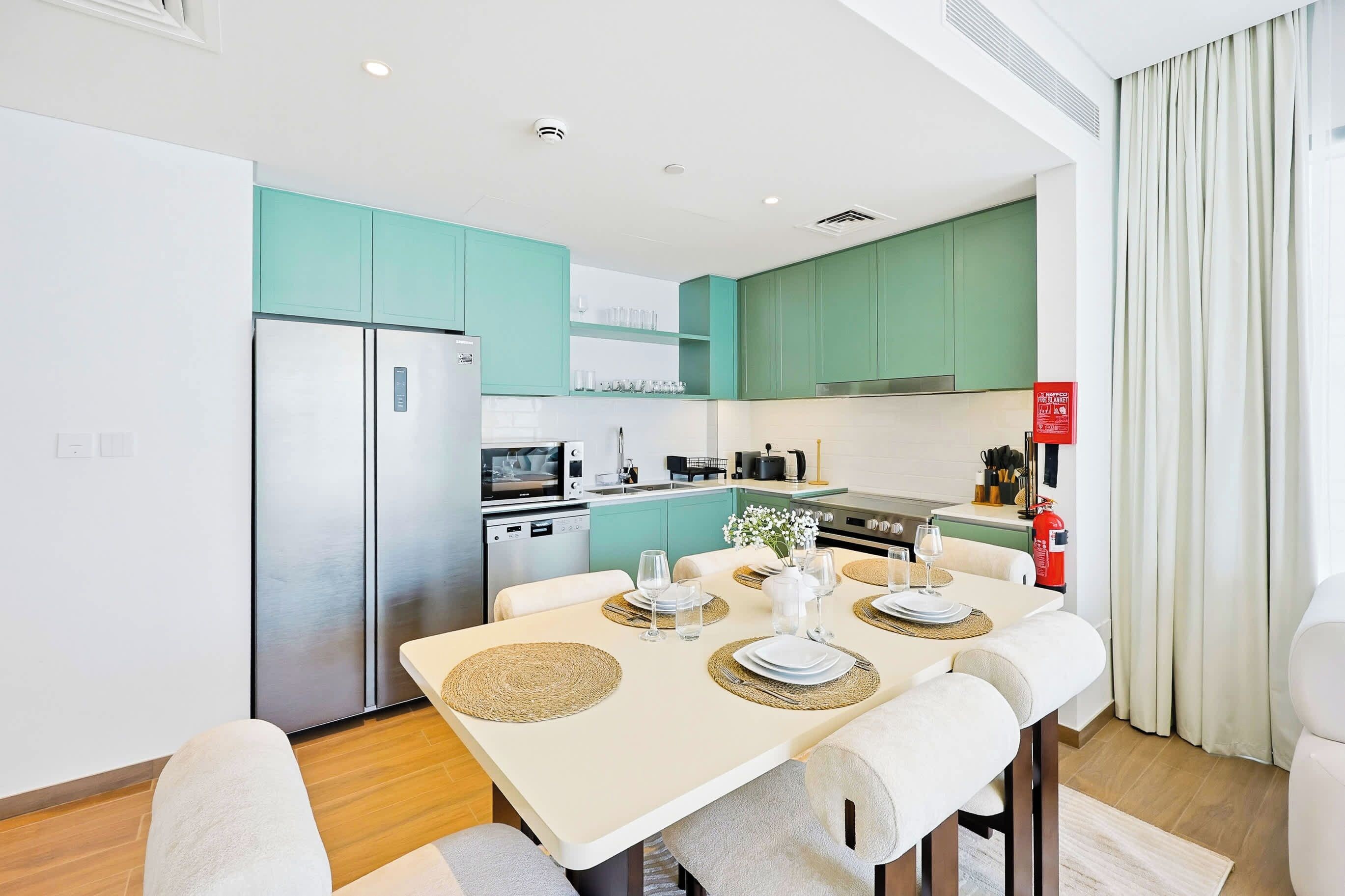 A modern kitchen with teal cabinets, a stainless steel refrigerator, and a dining area with a table set for two.